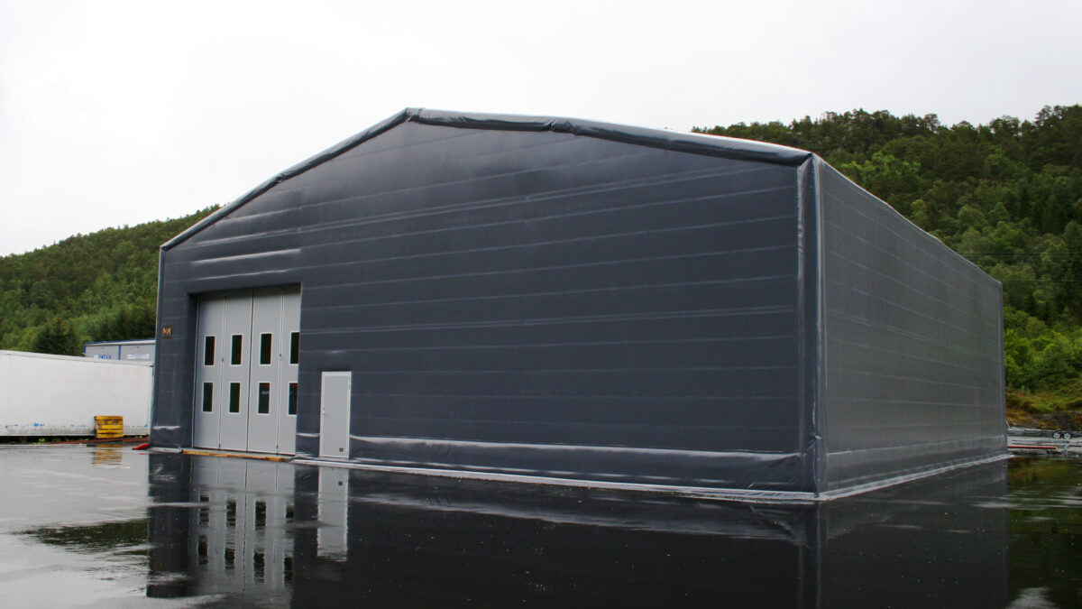 En grå isolert talthall oppført av Hallgruppen på en asfaltert overflate ved en grønn skog.